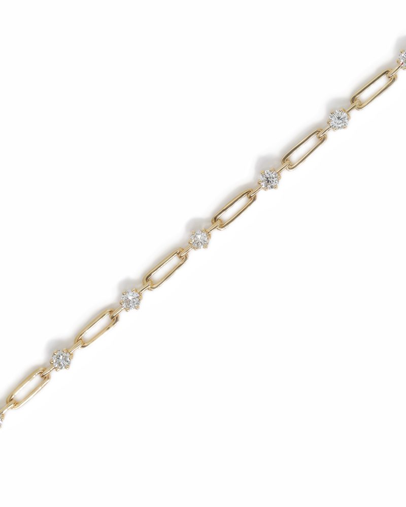 A yellow gold paperclip-link chain bracelet adorned with station diamonds. The thin bracelet is shown running diagonally across a white background. It has oblong links alternating with brilliant-cut round diamonds in prong settings. 一條黃金萬字夾鍊（Paperclip chain）風格的鑽石手鏈，呈對角線斜放於純白背景上。鍊節之間交替鑲嵌著亮麗的圓形切割鑽石。
