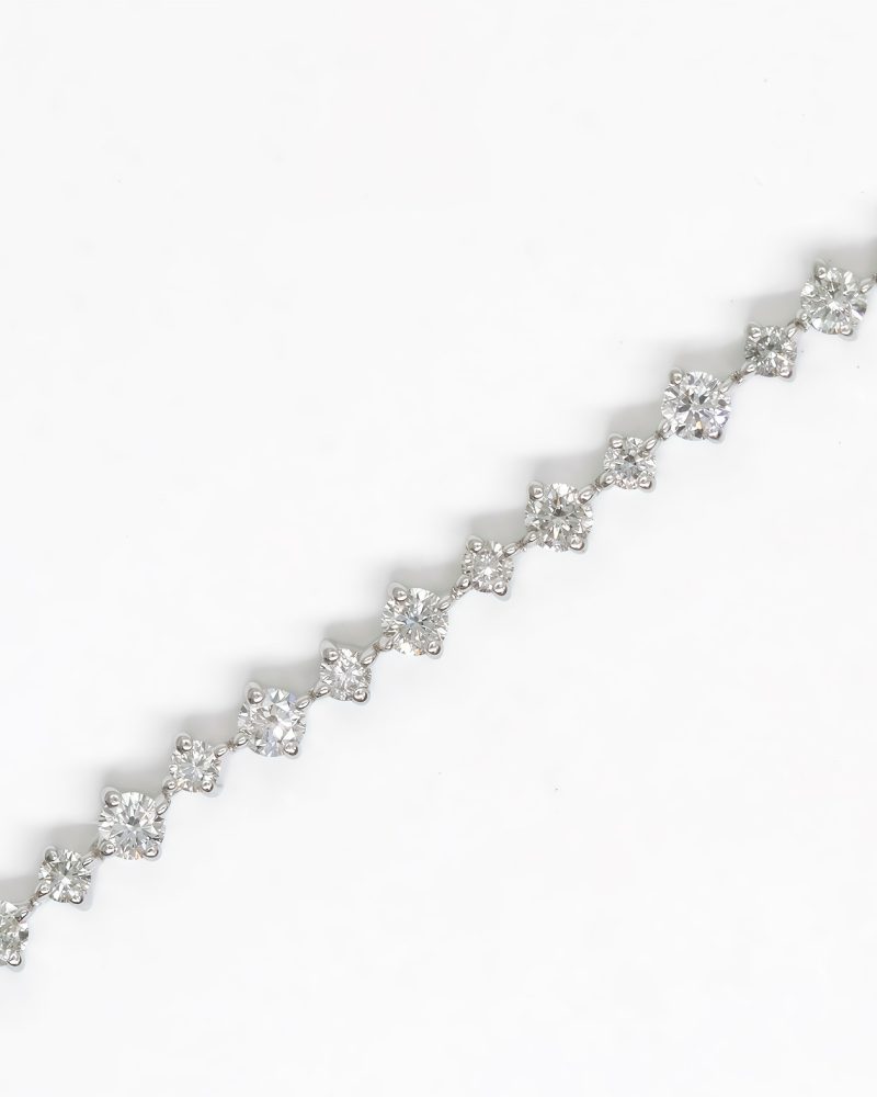 Close-up of a white gold diamond tennis bracelet featuring alternating large and small round brilliant diamonds laid diagonally against a white background. 白金鑽石網球手鍊特寫圖，展示斜向平鋪於白色背景上、大小交錯排列的圓形明亮式切割鑽石細節。