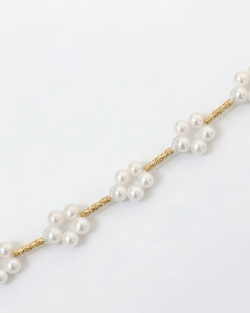 Close-up of a gold chain featuring cluster small white pearls formed into flower motifs interspersed with textured gold bar links against a white background. 黃金鍊飾細節圖，展示由小顆白色珍珠排列組成的花朵造型，並以帶有紋理的金屬管狀鏈節相連，背景為純白色。