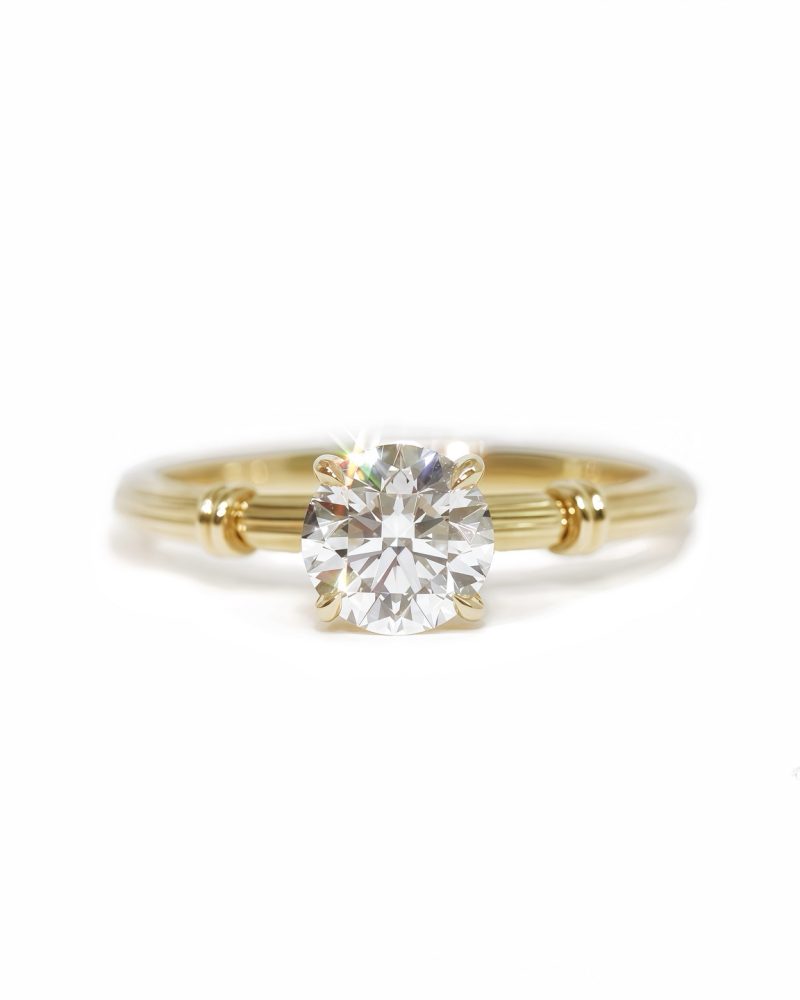 Front view of a yellow gold solitaire diamond engagement ring, featuring a round brilliant-cut diamond set on a textured band with roman column against a white background 黃金單鑽求婚戒指正面圖，展示中央爪鑲的圓形明亮式切割鑽石，圓形明亮式切割鑽石鑲嵌在帶有羅馬柱圖案的紋理戒圈上，背景為純白色。