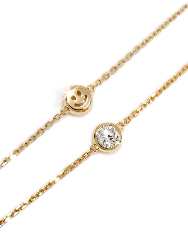 Two K yellow gold chain bracelets displayed diagonally against a white background, one featuring a polished smiley face charm and the other a round bezel-set solitaire diamond. K黃金細手鍊特寫圖，斜向展示兩條細緻金鍊：一條綴有亮面微笑臉孔（Smiley face）小吊飾，另一條綴有包鑲圓形單鑽，背景為純白色。
