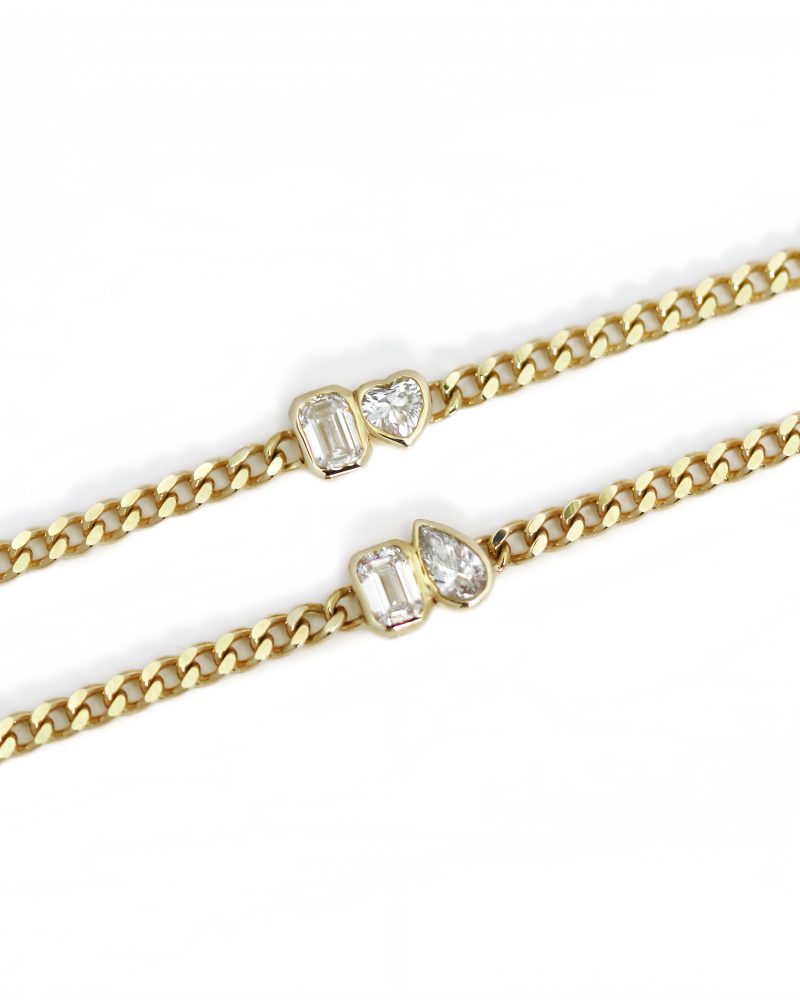 Two yellow gold cuban chain bracelets displayed diagonally against a white background, each featuring a bezel-set two-stone element: one with an emerald-cut and heart-cut diamond, and the other with an emerald-cut and pear-cut diamond. K黃金古巴古巴鍊手鍊特寫圖，斜向展示兩條手鍊：兩者皆包含祖母綠切工鑽石，分別搭配心形切工鑽石與梨形切工鑽石雙石包鑲配飾，背景為純白色。 Close-up view of a yellow
