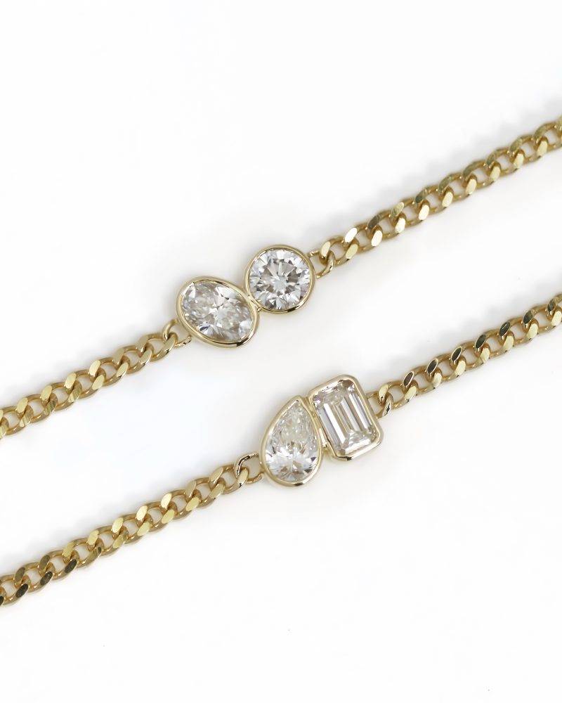 Two yellow gold cuban chain bracelets displayed diagonally against a white background, each featuring a bezel-set two-stone element: one with an emerald-cut and heart-cut diamond, and the other with an emerald-cut and pear-cut diamond. K黃金古巴古巴鍊手鍊特寫圖，斜向展示兩條手鍊：兩者皆包含祖母綠切工鑽石，分別搭配心形切工鑽石與梨形切工鑽石雙石包鑲配飾，背景為純白色。